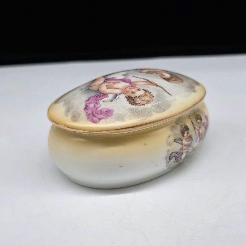May include: Oval porcelain trinket box with a lid. The lid features a painted scene of cherubs with pink and purple accents. The box has a cream and yellow gradient color scheme, with additional cherub figures painted on the side.