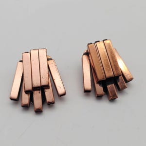 May include: A pair of copper-colored earrings. Each earring features a cluster of rectangular bars arranged in a fan-like design. The earrings have a vintage or retro aesthetic.