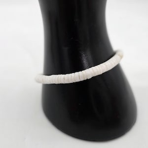 Puede incluir: Una pulsera de cuentas blancas se muestra sobre un soporte negro. La pulsera está hecha de pequeñas cuentas blancas redondas ensartadas. Las cuentas parecen uniformes en tamaño y forma, creando un diseño simple y elegante. La pulsera es una delicada pieza de joyería.