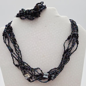 May include: A multi-strand necklace and bracelet set. The necklace features strands of small, iridescent beads with dark wooden accents and square, iridescent beads. The bracelet mirrors the necklace design. The jewelry is displayed on a white mannequin.