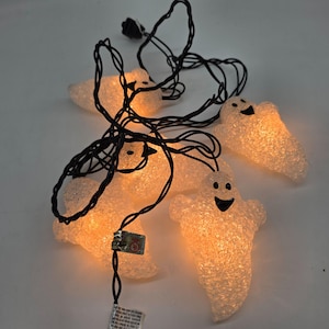 May include: String of Halloween ghost lights. Each ghost is white and has a smiling face. The ghosts are lit with a warm, orange glow. The string has a black cord and a standard plug.