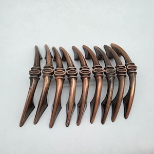 Puede incluir: Un conjunto de nueve tiradores de armario de color bronce. Cada tirador tiene un asa curvada con un detalle ovalado decorativo en el centro. Los tiradores están dispuestos en fila, mostrando su diseño y acabado. Adecuado para muebles o armarios.