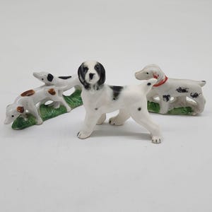 May include: Three porcelain figurines of dogs. Two of the dogs are white with black spots and are standing on a green base. The third dog is white with black spots and a red collar and is standing on a green base.