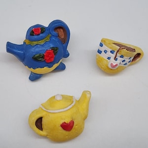 Puede incluir: Tres miniaturas de cerámica con temática de té. Una tetera azul con detalles de rosas rojas y ribete amarillo, una taza de té amarilla con una banda estampada azul y blanca y una tetera amarilla con un corazón rojo.