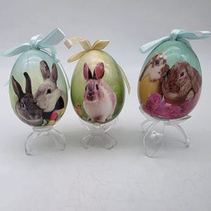 May include: Three decorative Easter eggs with bunny illustrations. Each egg is oval-shaped, resting on a clear plastic stand, and adorned with a pastel ribbon. The eggs feature images of rabbits in various poses, set against a soft, green background.