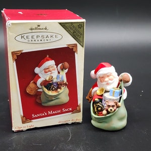 Könnte beinhalten: Ein Hallmark Keepsake Ornament mit dem Weihnachtsmann, der einen grünen Sack voller Spielzeug hält. Das Ornament ist mit "Santa's Magic Sack" und "2005 VIP Gift" beschriftet.