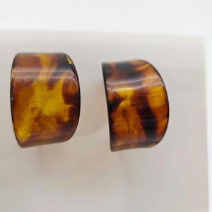 May include: A pair of tortoise shell-patterned earrings. The earrings are a half-circle shape with a glossy finish. The colors include shades of brown and amber.