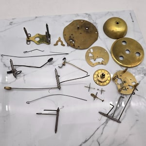 May include: A collection of vintage clock parts, including brass gears, plates, springs, and levers. The components are scattered on a white surface, showcasing the intricate details of the clock mechanism. The parts vary in size and shape, with a mix of metallic finishes.