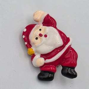 May include: A glossy Santa Claus brooch featuring a red suit with white trim, a red hat with white polka dots and a yellow pom-pom, a white beard, and black boots. The figure is depicted in a dynamic pose with one arm raised and one arm extended.