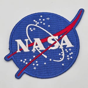 May include: A circular embroidered patch featuring the blue NASA logo. The design includes white text reading NASA, a white orbital path, a red swooping vector line, and small white star-like dots scattered across a deep blue background.
