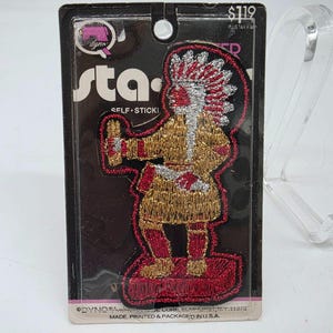 Puede incluir: Un parche autoadhesivo bordado que representa una figura nativa americana en hilo dorado, rojo y plateado. El parche está sobre un respaldo negro con la palabra "sta" y "SELF STICKI" impresas. El parche está empaquetado en un respaldo de cartón.