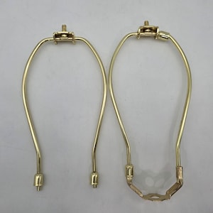 Puede incluir: Dos arpas de lámpara doradas. Las arpas tienen un diseño curvo en forma de U con una parte superior e inferior decorativas. Las arpas están hechas de metal y están diseñadas para sujetar una pantalla de lámpara en su lugar. Las arpas son para una lámpara.