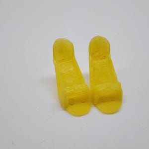 以下が含まれることがあります： 白い背景に、靴の中敷きやサポートのような、2つの小さな黄色のプラスチック製の物体が並んでいます。それぞれ丸みを帯びた上部と、わずかにテクスチャ加工された表面を持つ長方形のベースがあります。