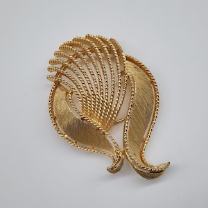 Könnte beinhalten: Goldfarbene Vintage-Brosche im Blätterdesign. Die Brosche hat zwei große strukturierte Blätter und ein zentrales fächerförmiges Element mit parallelen Linien. Die Ränder sind mit einer gedrehten, seilartigen Textur umrandet, was ein detailliertes Accessoire ergibt.
