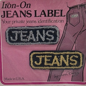 May include: Iron-on jeans labels with the word "JEANS" embroidered in white and gold on a black background. The labels are rectangular and have a stitched border. The packaging is pink and includes the text "Made in U.S.A."