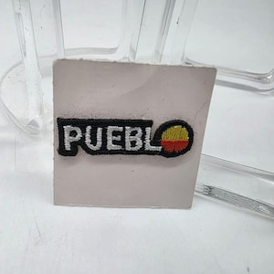May include: Embroidered patch with the word "PUEBLO" in white letters on a black background. The patch includes a circular design with yellow, red, and orange colors. The patch is on a light pink surface.