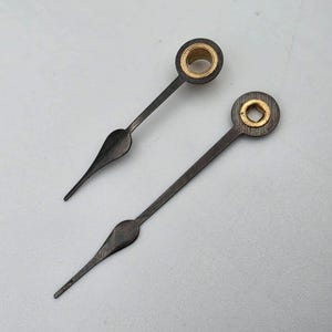 May include: Two dark gray clock hands with a metallic finish, each featuring a pointed tip and a circular base. The base of each hand contains a gold-colored center insert, with one showing a circular opening and the other a square opening.