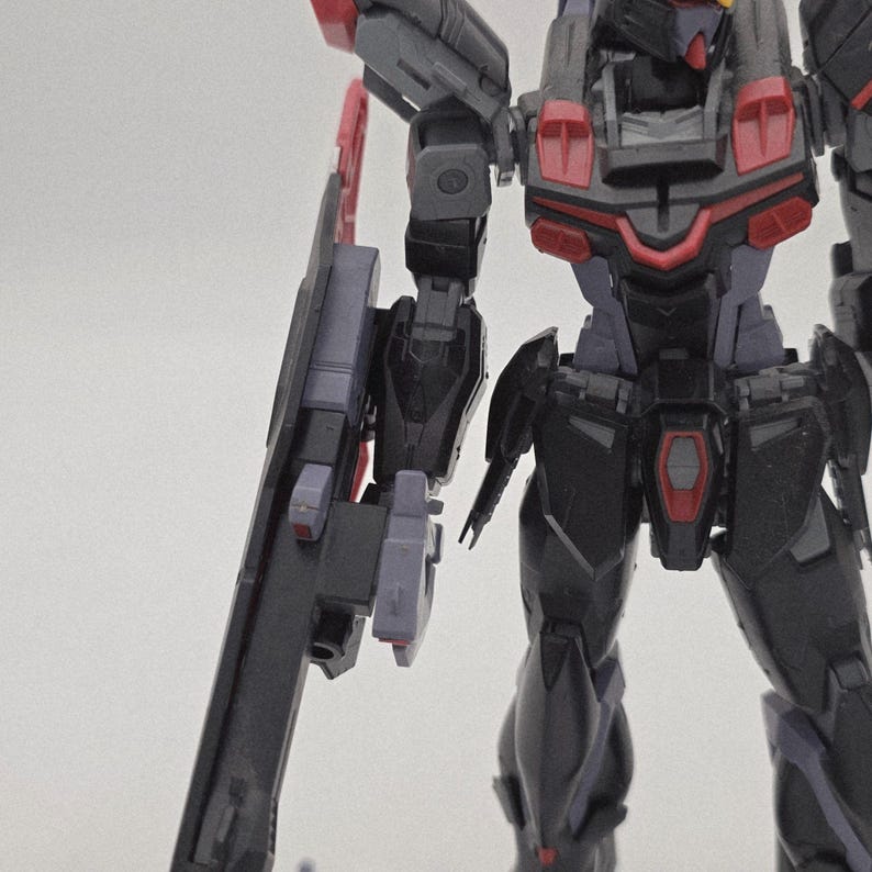 Peut inclure: Maquette de robot partiellement assembl&eacute;e, avec des &eacute;l&eacute;ments noirs, gris et rouges. Le kit comprend une grande arme lat&eacute;rale. Le mod&egrave;le pr&eacute;sente des d&eacute;tails m&eacute;caniques complexes, typiques des maquettes, avec une attention particuli&egrave;re &agrave; la pr&eacute;cision.