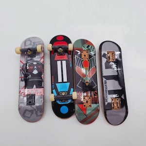 May include: Four miniature skateboards with various designs. One features a ninja graphic, another has a geometric pattern, and a third displays a red and green design. The fourth has the word "Skateboards" printed on it. Each has small wheels.