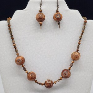 May include: A necklace and earring set featuring large, round, brown beads with a circular design. The necklace has multiple beads and smaller accent beads. The earrings have a similar design, with a hook closure. The jewelry is displayed on a white mannequin.