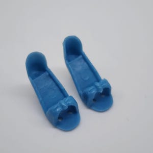 以下が含まれることがあります： 鮮やかな青色の人形の靴のペア。各靴はつま先にリボンのディテールと小さな開口部があります。靴は丈夫で滑らかな素材でできており、小さな人形に合うように設計されています。