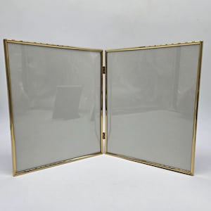 May include: A hinged, double-sided photo frame with a gold-colored metal frame. The frame is open, revealing two gray-toned spaces for photos. The frame is designed to stand upright and is suitable for displaying two pictures.