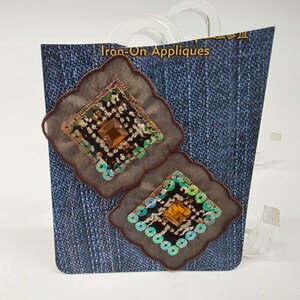May include: Two iron-on appliqués with a diamond design. Each appliqué has a brown border, a central square with a brown gem, and is decorated with green sequins. The background is a denim print, and the package reads "Iron-On Appliques."