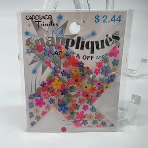 Può includere: Una confezione di plastica trasparente contenente un'applicazione Snappliques a motivi floreali. L'applicazione ha la forma di una girandola con una varietà di fiori colorati. La confezione riporta la scritta "CAROLACE Trimtex" e "Snappliques".