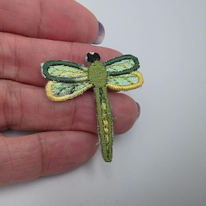 May include: An embroidered dragonfly patch featuring a green body with yellow and light green wing details. The insect design has a black head and a segmented tail, with intricate stitching throughout the wings and body for texture.