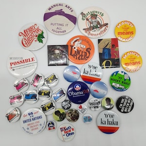 May include: A collection of vintage pinback buttons with various designs and text. Some buttons feature phrases like "Merry Christmas," "Mission Possible," and "Obama '08." Other buttons have graphics and text related to movies, organizations, and events.