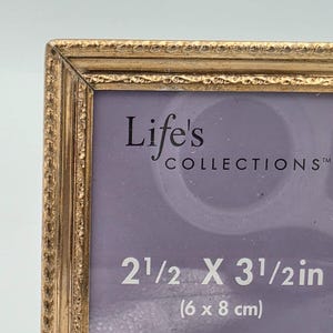 May include: A gold-toned picture frame with a textured border. The insert displays the text Life's COLLECTIONS in black, followed by the dimensions 2 1/2 X 3 1/2 in (6 x 8 cm) in white, set against a light purple background.