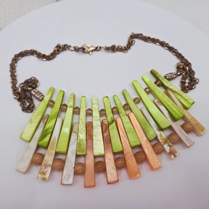 May include: A statement necklace featuring a bronze-colored chain and a fan-shaped pendant. The pendant is composed of green, orange, and white shell-like pieces, interspersed with small, round beads. The necklace has a lobster clasp closure.