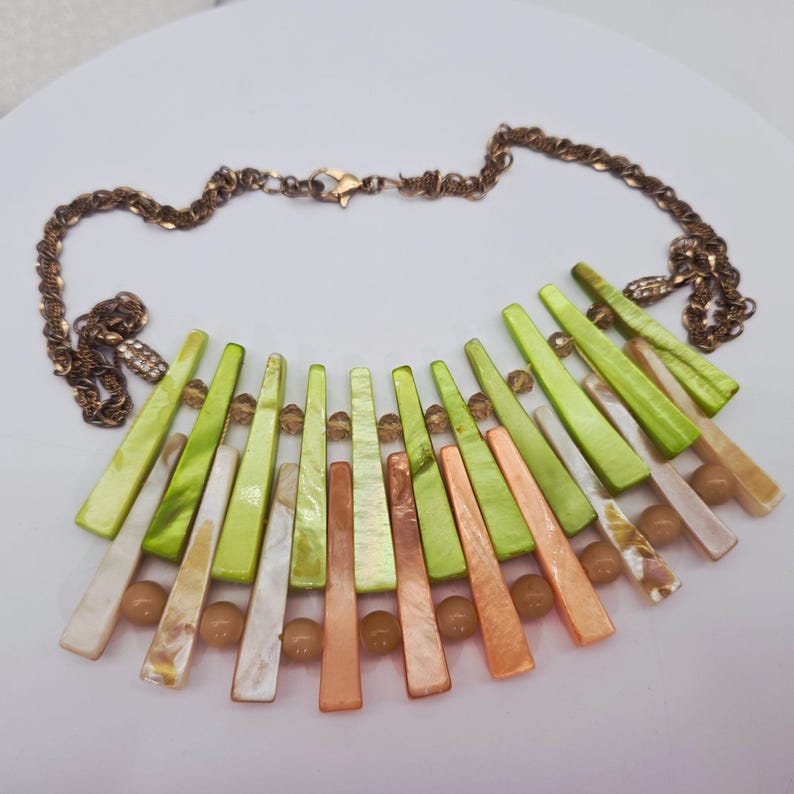 May include: A statement necklace featuring a bronze-colored chain and a fan-shaped pendant. The pendant is composed of green, orange, and white shell-like pieces, interspersed with small, round beads. The necklace has a lobster clasp closure.