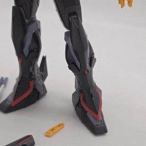 Peut inclure: Gros plan sur les jambes et les pieds d'une figurine d'action noire et rouge. La figurine a des accents rouges et violets. Une petite pi&egrave;ce jaune et une pi&egrave;ce noire sont au premier plan.