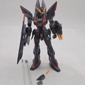 Peut inclure: Maquette d&eacute;taill&eacute;e d'un robot m&eacute;cha noir et rouge avec des accents gris et jaunes. Le robot a une grande arme sur le c&ocirc;t&eacute; gauche et plusieurs pi&egrave;ces d&eacute;tach&eacute;es. Le mod&egrave;le est debout sur une surface blanche.