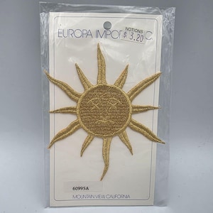 Puede incluir: Un parche bordado dorado con forma de sol y un rostro con ojos cerrados y boca curva. El parche está montado sobre una tarjeta de soporte rectangular blanca con la etiqueta EUROPA IMPORT INC., el texto 60995A y MOUNTAIN VIEW CALIFORNIA.