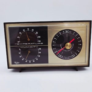May include: A vintage Taylor combination weather instrument. The device features a temperature and humidity gauge on the left and a barometer on the right. The face is black and gold, with a dark brown frame and base.