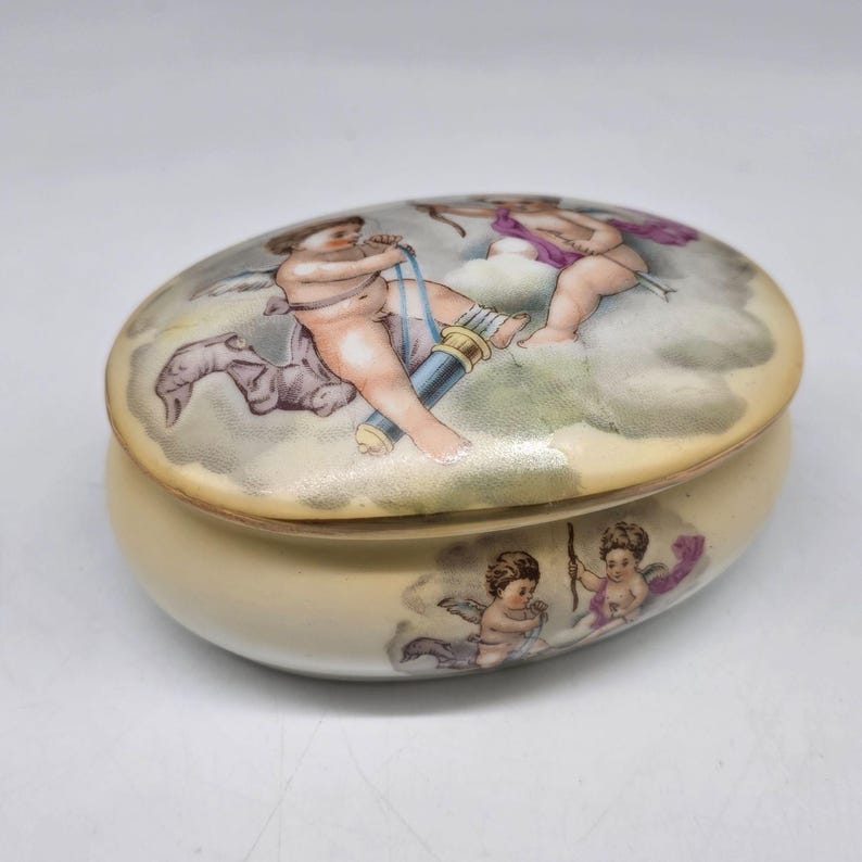 May include: Oval-shaped trinket box with a cream-colored base and lid. The lid features a painted scene of cherubs with bows and arrows, set against a cloudy background. The box has a glossy finish and a decorative design.