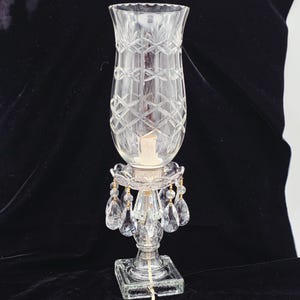 Puede incluir: Una lámpara de huracán de vidrio transparente con un patrón de diamantes, una base cuadrada y detalles de cristal colgantes. La lámpara tiene una vela dentro y mide aproximadamente 30 cm de alto.