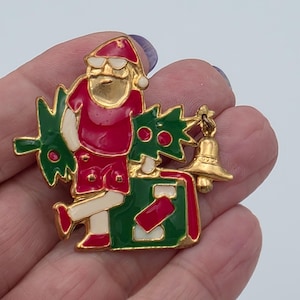 Pode incluir: Um broche de metal dourado com uma figura esmaltada vestindo um chapéu vermelho, camisa vermelha e calções brancos, carregando uma árvore de Natal verde. A figura está ao lado de uma mala verde com detalhes vermelhos e um pequeno sino dourado.