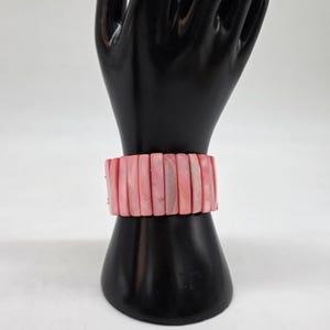 May include: A pink bracelet composed of rectangular, polished beads. The bracelet is displayed on a black mannequin hand against a white background. The beads have a subtle, natural variation in color, adding depth to the design.