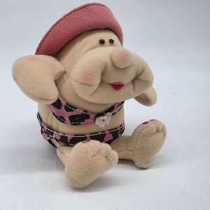 May include: A plush toy figure with a pink hat, a pink and black leopard print bikini, and a large nose. The figure has rosy cheeks and red lips. The toy is sitting down with its arms and legs outstretched.