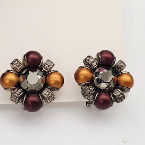 May include: Pair of vintage clip-on earrings. Each earring features a central faceted gray stone surrounded by alternating burgundy and gold-colored orbs. The earrings have a silver-tone metal floral design.