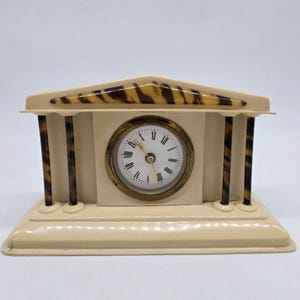May include: A vintage ivory-colored clock with a decorative architectural design. The clock features a round face with Roman numerals, a gold-tone bezel, and brown and tan accents. The clock is approximately 6 inches wide and 4 inches tall.