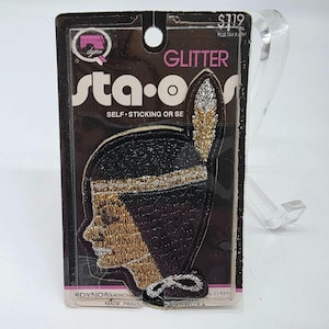 Puede incluir: Un parche de purpurina autoadhesivo que representa el perfil de una mujer nativa americana con un tocado de plumas. El parche es negro, dorado y plateado, con el texto "sta-o" y "GLITTER" visibles en el embalaje.