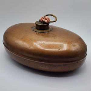 May include: An oval-shaped copper hot water bottle with a metallic finish. The item features a central brass screw cap with an attached metal ring and a small piece of frayed, light-coloured fabric tied through the ring for handling.