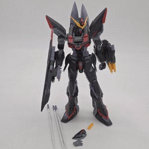 Peut inclure: Maquette d&eacute;taill&eacute;e d'un robot noir et gris avec des accents rouges et jaunes. Le robot a de grandes &eacute;paulettes, une arme sur le c&ocirc;t&eacute; gauche et plusieurs pi&egrave;ces d&eacute;tach&eacute;es. Le mod&egrave;le est pos&eacute; sur une surface blanche.