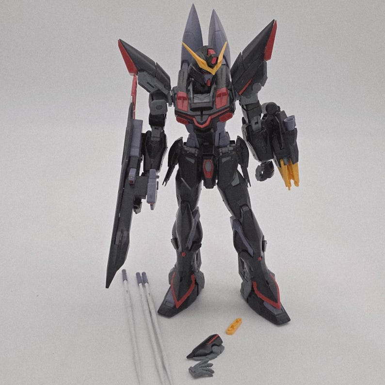 Peut inclure: Maquette d&eacute;taill&eacute;e d'un robot noir et gris avec des accents rouges et jaunes. Le robot a de grandes &eacute;paulettes, une arme sur le c&ocirc;t&eacute; gauche et plusieurs pi&egrave;ces d&eacute;tach&eacute;es. Le mod&egrave;le est pos&eacute; sur une surface blanche.
