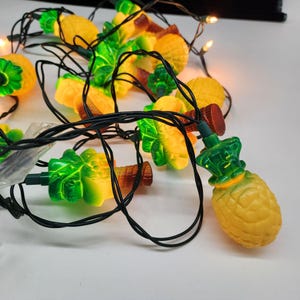May include: String of novelty lights featuring yellow pineapple and green palm tree shapes. The lights are strung on a black wire and have a warm glow. Perfect for adding a tropical touch to any space.