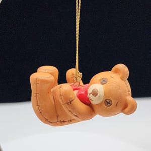 Può includere: Un ornamento di orsacchiotto di ceramica marrone con un fiocco rosso appeso a uno spago dorato. L'orso è sdraiato sulla schiena con le zampe in aria.